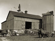 Two men with horses, buggy and farm implements in front of large bank barn.