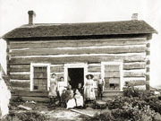Two women and seven children in front of single-storey squared timber cabin.