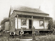 Two elderly men in front of 1½-storey frame house with decorative pump in foreground.
